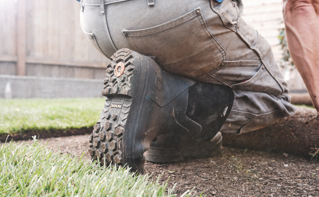 Image features a man laying sod in a short sleeve shirt and Bogs boots. 